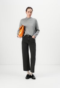 Grau gestrickter Pullover mit hohem Kragen. Schwarze weit geschnittene Jeans mit rohem Saum. Orangefarbene Clutch-Tasche mit glatter Textur. Schwarze Loafers, die im Kontrast zu beigefarbenen Socken stehen.
