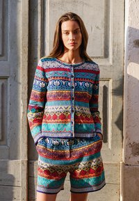 Naine seisab käed taskus, kandes värvilist mustrilist pika varrukaga pluusi ja sobivaid lühikesi pükse rustikaalse puidust ukse taustal.