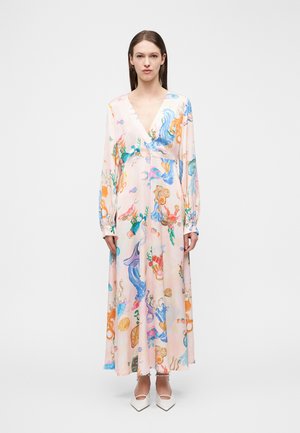 Kobieta w różowej maxi sukience z długim rękawem, w pastelowym odcieniu, ozdobionej kolorowymi, abstrakcyjnymi wzorami inspirowanymi oceanem, nosząca białe szpiczaste buty na jednolitym białym tle.