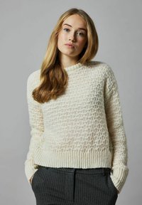 Cremiger Strickpullover mit einem strukturierten Rautenmuster und gerippten Bündchen. Kombiniert mit dunklen, gestreiften Hosen. Glatte, warme Textur.