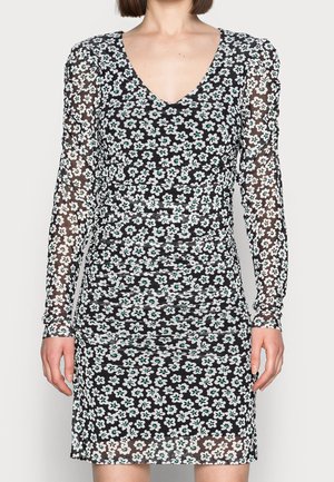 Kobieta ubrana w czarną mini sukienkę z długim rękawem, z białym i turkusowym kwiatowym wzorem oraz marszczonym gorsetem, stojąca na jednolitym jasnym tle.