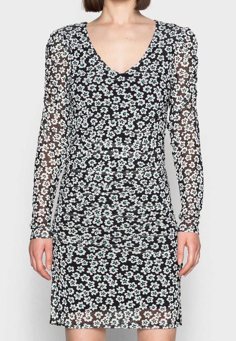 Femme portant une mini-robe à manches longues noire avec un motif floral blanc et turquoise et un corsage froncé, debout devant un fond clair uni.