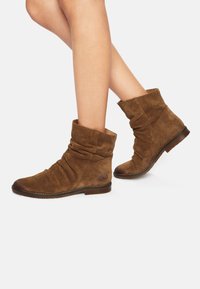 Botines de ante marrón con un diseño arrugado, puntera redondeada, tacón bajo y detalles de costura. Presenta un logo en el lado exterior.