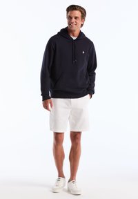 Sweatshirt à capuche bleu marine avec poche kangourou, arborant un petit logo sur la poitrine. Associé à un short blanc et des baskets blanches.