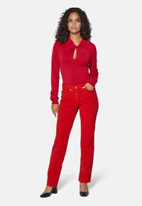 Rotes Langarmoberteil mit einem Schlitzdetail, kombiniert mit roten geraden Jeans und schwarzen Absatzschuhen. Glatte Textur, taillierte Passform.