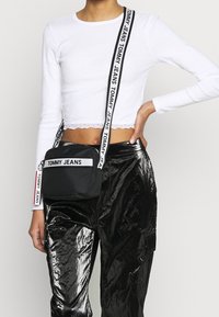 Black fabric crossbody bag with a white logo strap, featuring a rectangular shape and zipper closure, displayed with shiny, textured black pants.