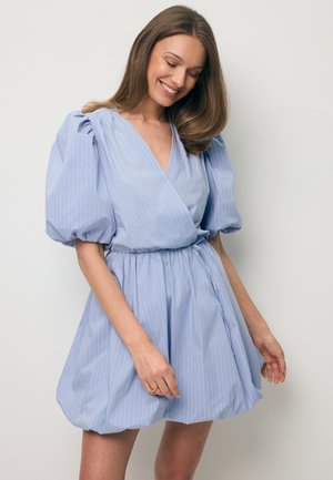 Lachende vrouw met lang bruin haar, die een lichtblauwe wikkeljurk met pofmouwen en subtiele witte strepen draagt, staand tegen een effen achtergrond.