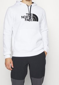 Persona vestida con una sudadera blanca con el logo negro de "The North Face" y pantalones negros con parches grises en las rodillas, de pie con una mano en el bolsillo.
