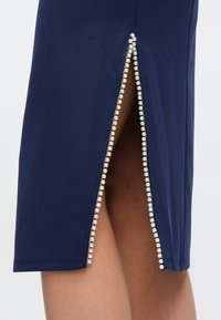 Φούστα μολύβι σε ναυτικό μπλε με πλαϊνή σχισμή διακοσμημένη με σειρές από μικρές πέρλες κατά μήκος της ραφής. Ομαλή υφή υφάσματος, εφαρμοστό σχέδιο.