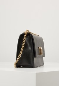 Borsa a tracolla in pelle nera con una forma strutturata, tracolla in catena dorata e pattina frontale con un dettaglio di chiusura dorata. Dettaglio della superficie testurizzata.