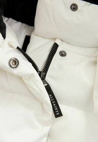Vit puffjacka med svart dragkedja och en märkesetikett. Slät struktur med silverfärgade knappar som visar en lager-design.