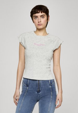 Jeune personne aux cheveux courts et foncés portant un T-shirt gris ajusté à manches courtes avec le texte rose "Ocean Drive Miami" et un jean bleu taille haute.
