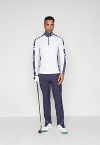 Chemise de sport blanche à manches longues avec des manches à motif marine, associée à un pantalon marine et des chaussures de golf blanches, tenant un club de golf.