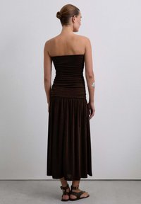Vestido marrón sin tirantes con textura fruncida y una falda larga y fluida hasta los tobillos. Llevado con sandalias negras con tachuelas. Sin patrones visibles ni acentos.