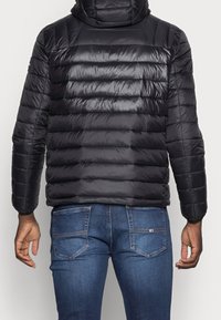 Zwarte donzen jas met gestikte horizontale panelen, hoge kraag en lichtgewicht glanzende stof. Gecombineerd met blauwe spijkerbroek.