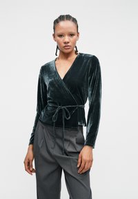 Tummanvihreä samettinen kääntötoppi, jossa on pitkät hihat, syvä V-pääntie ja sidottu vyöyksitys. Yhdistetty vaaleanharmaisiin laskostettuihin housuihin.