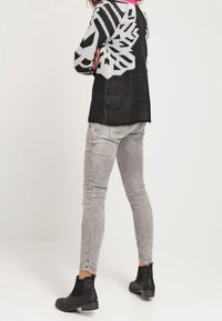 Henkilö, jolla on päällä musta-valkoinen kuvioitu villapaita, vaaleanharmaat skinny-farkut ja mustat nilkkurit, seisoo käsivarsia ristissä.