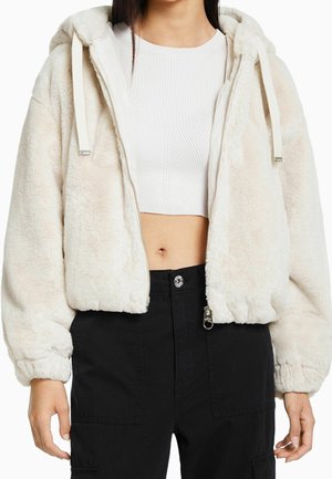Kvinde iført en hvid kort ribbet top og en beige fluffy lynlåstrøje med sorte bukser med høj talje.