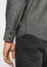 Primo piano della mano sinistra di una persona accanto a un polsino di una camicia grigia a maniche lunghe con bottoni e la tasca di un jeans neri.
