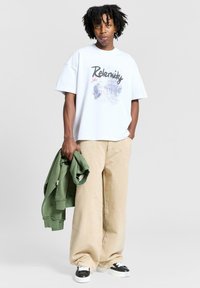 Valkoinen t-paita, jossa on "Relemity" -grafiikka, beiget leveät housut, vihreä neuletakki kädessä, mustavalkoiset sneakersit, rento asu.