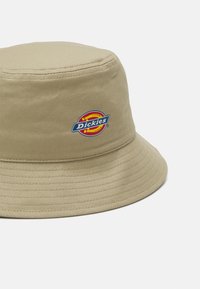 Beige színű pamut vödrös kalap lapos koronával és széles karimával. Elöl egy kerek logó folt található kék színű felirattal.