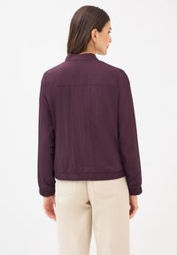 Een vrouw in een paarse jas en beige broek staat met haar rug naar de camera, waardoor de outfit wordt getoond tegen een simpele witte achtergrond.