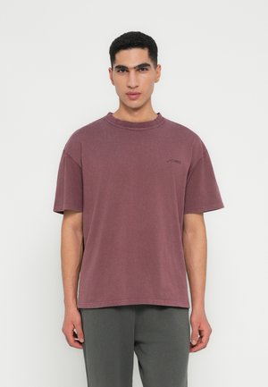 Ung mand med kort krøllet hår iført en vasket, oversize bordeaux T-shirt og mørkegrå bukser, stående mod en ensfarvet baggrund.