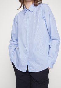 Personne portant une chemise à rayures bleu clair à boutons avec manches longues et pantalon noir, mains dans les poches, fond blanc.