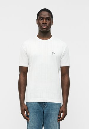 Homme portant une chemise blanche à manches courtes avec de fines rayures verticales et un petit logo brodé sur la poitrine, associée à un jean bleu.