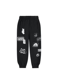 Pantalons de jogging noirs avec taille et poignets élastiques, présentant divers imprimés graphiques blancs et patchs, y compris des symboles de paix et des thèmes spatiaux.