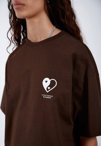 Camiseta de algodón marrón con un corte holgado, que presenta un logo de corazón blanco en el pecho y una pequeña etiqueta de la marca debajo.