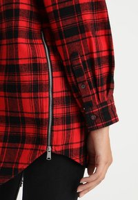 Czerwona i czarna koszula flanelowa w kratę z zamkiem po boku. Posiada guzikowe mankiety oraz zaokrąglony dół, co nadaje nowoczesny kształt.