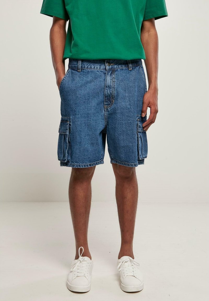 Homme portant un short cargo en denim bleu, un t-shirt vert et des baskets blanches, debout les mains dans les poches devant un fond uni.