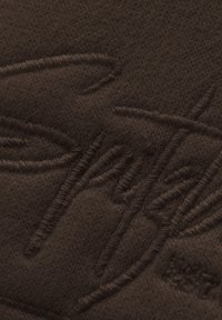 Braun gestrickter Stoff mit erhabener Stickerei eines Logos. Die Textur ist durchgehend glatt und einheitlich, ohne zusätzliche Muster oder Akzente.