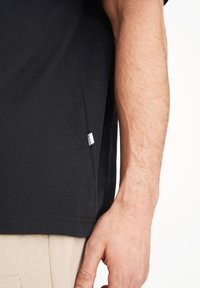 Camiseta de algodón negra, de manga corta, con etiqueta en la costura lateral. Textura suave, diseño simple, costura visible a lo largo del dobladillo. Brazo parcialmente visible.