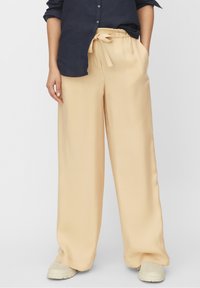 Persona che indossa pantaloni beige a gamba larga con vita regolabile, scarpe beige chiare e una maglietta a maniche lunghe blu scuro con le maniche arrotolate.