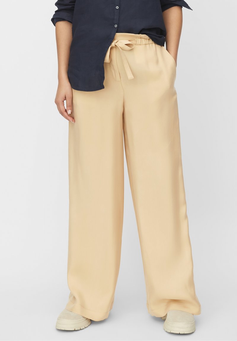 Persona che indossa pantaloni beige a gamba larga con vita regolabile, scarpe beige chiare e una maglietta a maniche lunghe blu scuro con le maniche arrotolate.