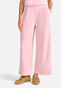 Pantaloni a gamba larga rosa realizzati in tessuto morbido, dotati di due tasche frontali e orlo ripiegato. Abbinati a scarpe con lacci color marrone chiaro.
