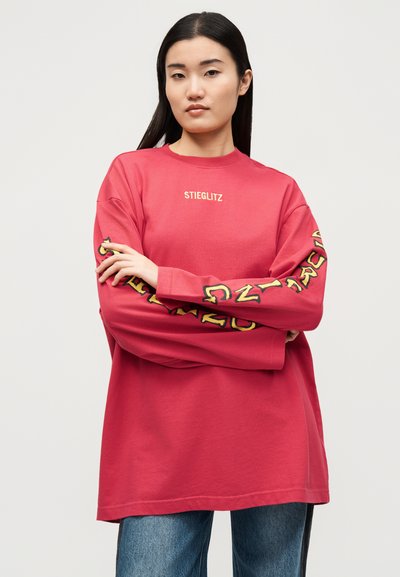 Νεαρή γυναίκα με μακριά σκουρόχρωμα μαλλιά φοράει μια υπερμεγέθη κόκκινη μπλούζα με τη λέξη "STIEGLITZ" στο στήθος και γραφικά γράμματα στα μανίκια, συνδυασμένη με τζιν.