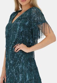 Turkusowa sukienka z frędzlami, charakteryzująca się głębokim dekoltem w kształcie litery V oraz misternym zdobieniem koralikami. Światło odbija się od cekinów, podkreślając fakturę i wzór.