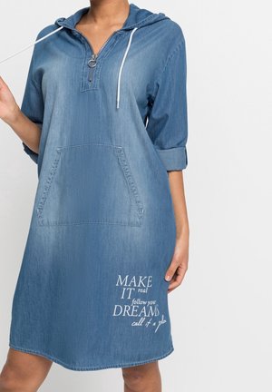 Woman wearing blue denim hooded dress with front pocket and white text reading "Make it real, follow your dreams, call it a plan" on the skirt.