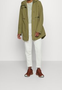 Oliwkowa wodoodporna kurtka z kapturem, z dwiema kieszeniami na klapki, zestawiona z białymi dresami i brązowym obuwiem sportowym. Gładkie wykończenie materiału.