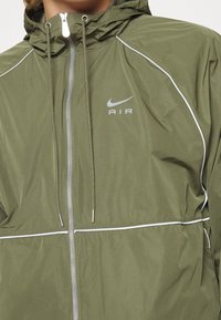 Coupe-vent Nike vert olive avec des accents argentés, capuche et fermeture éclair à l'avant. Présente des passepoils blancs et un logo sur la poitrine. Texture lisse.