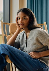 Gray knitted cardigan with three buttons, paired with blue denim jeans. Seated on a wooden chair against a backdrop of textured blue curtains.