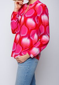 Lyserød langærmet bluse med et abstrakt mønster i lysere rosa og røde nuancer. Har en V-halsudskæring og løs pasform, kombineret med blå jeans.