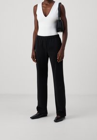 Vit ärmlös bodysuit med djup V-ringning, kombinerad med svarta byxor med elastisk midja. En minimalistisk svart handväska och platta svarta skor fulländar outfiten.