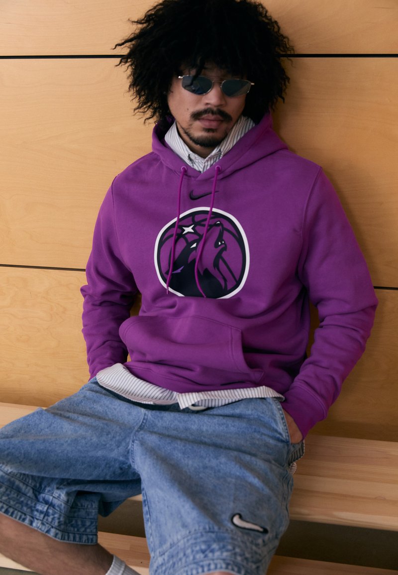 Sweatshirt roxa com capuz e logo de um leão, combinado com calções de ganga claros. O modelo está sentado num banco de madeira com um fundo de madeira.