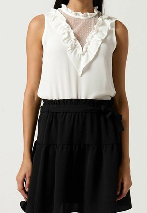 Femme portant un chemisier blanc sans manches avec un détail volanté en maille en V et une jupe noire taille haute nouée à la taille avec un nœud.
