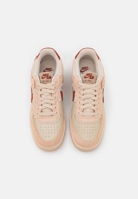 Nike Air Force 1 sneakers in light pink and beige suede with red accents. Features perforated toe, laces, and branded tongue. Smooth texture.