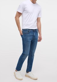 Pánské modré džíny se zúženým střihem, s bočními kapsami a decentními bledými odstíny, kombinované s bílým tričkem a bílými teniskami.